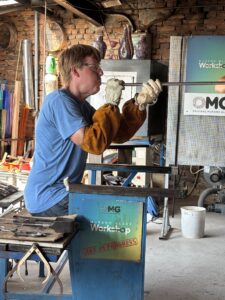 Carl wearing a blue t-shirt, jeans, white leather gloves, and brown leather protective forearm covers. He is sitting on a bench and you see him from the side as he holds a glass blowing tube and blows into it. Behind him you can see parts of a glass-making workshop.