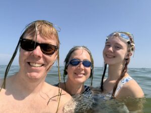 Carl with sunglasses on and seaweed in his hair, Jenny with goggles, Amy with goggles on top of her head, all submerged in the ocean with their heads above water