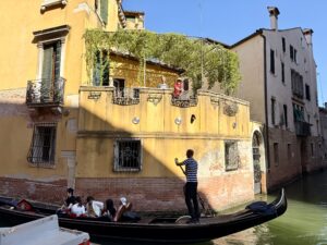 A yellow three-story building mostly in the sun. The second-story has a terrace with green vines. Sarah is wearing a red t-shirt and is standing on the terrace waving. Jenny is in a dark blue t-shirt next to her but is harder to see.  A gondolier is rowing past, perfectly centered 