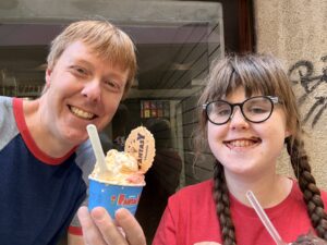Carl and Sarah smiling while holding cups of gelato with spoons sticking out of the gelato. There are cookie wafers sticking out of the gelatos too. Sarah has some chocolate around her mouth. She is wearing black and white striped glasses. She has a red t-shirt. Carl has on a blue t-shirt