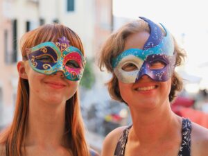 Close-up of Amy and Jenny outside with a blurred background of Venice. They are wearing Venetian masks that they painted. Amy’s mask is purple, pink, and turquoise with gold decoration. Jenny’s is silver, purple, and turquoise with silver decoration. 
