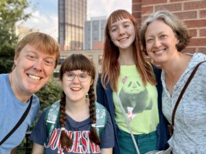 Carl, Sarah, Amy, and Jenny are outside, clustered together and smiling with brick buildings behind them. The sun is shining. Carl has a blue t-shirt on. Sarah is wearing a dark blue t-shirt with "Duquesne" printed in red letters across it. Amy is wearing a light green t-shirt with a gray panda on it. Jenny has on a light gray long-sleeved shirt. 
