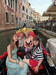 In a gondola in a canal in Venice with red brick buildings on either side. Amy is in a sea green sun dress. Sarah is wearing a black and white horizontal striped shirt and red shorts. Behind the girls, Carl and Jenny are also seated. Carl is in a grey polo shirt and navy blue chinos. Jenny is in a multicolored flowered sundress. 