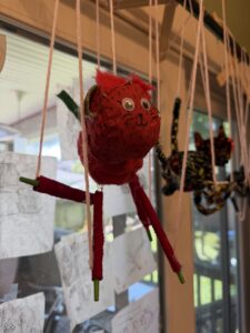 Close up of a red cat puppet that looks like a strawberry turned into a cat. It has strings attached to each leg and it suspended from cross bars above it.