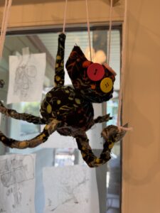 A black cat puppet with multi-colored musical notes as part of the black fabric. The cat face has two large button eyes, one yellow and one red. The legs are attached to strings and the whole thing is suspended in the air, attached to cross-bars above it.