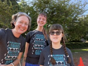 Jenny, Amy, and Sarah smile at the camera after the Run Around the Square. They are all wearing matching gray t-shirts that say "Run Around the Square" in teal print with white trim. Amy's face is flushed after the exertion of cartwheeling 1.5K. Sarah is tired. Sarah's glasses are tinted to sunglasses.
