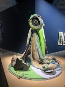 Art piece from the Biennale Arte 2026 in Venice. It looks like a large eye in a circle wearing a cape. The colors are white, brown, and green. 