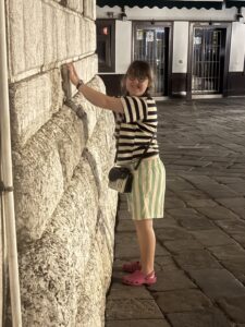 Sarah is wearing green and white striped shorts and a black and white striped shirt. She is seen from the side standing next to a stone bell tower and pretending to remove a stone, with her face turned to the camera and smiling. 