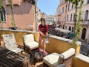 Sarah in red t-shirt and pink shorts standing on a low brown table in between two white-cushioned chairs on a terrace in the sunshine with a Venetian canal and buildings behind her. She is holding a small outside portable lamp thingie as a pretend oar and pretending to row a gondola.