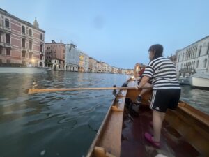 Sarah in a black and white striped t-shirt and black shorts with white stripes down the side, seen from behind as she looks to her left. She is holding an oar the sticks out to the left. She is standing in a batela that is floating on the Grand Canal in Venice. You can see buildings on either side of the canal. It is dusk.