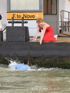 A woman in a red dress kneels by the side of a vaporetto stop in Venice with a yellow sign indicating it is the F.te Nove stop. She is reaching down to help a man in a blue long sleeved shirt climb out of the water. He fell in after doing a handstand on the black metal boat tie-up posts that are to the left of the photo, just above where he is in the water.