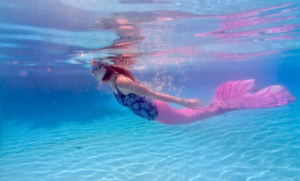 An underwater photo with Amy in a blue swimsuit top, blue goggles, and a large pink mermaid tail.