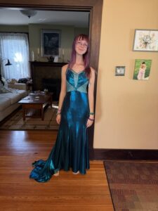A photo of Amy standing in a wide doorway, with a living room sofa and coffee table in the background. There are two art prints of Amy and Sarah on the wall next to Amy. Amy is wearing a shimmery, satiny dark turquoise blue evening gown with stitching and sequins that make it look like a mermaid tail shape at the top. Her arms are at her sides. Her purple hair is hanging straight down to her shoulders. She is smiling with her lips closed.