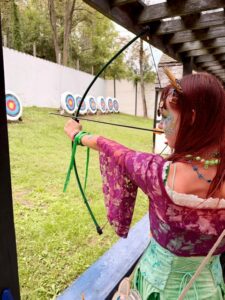 A photo of Amy as she faces away from the camera holding a bow and arrow, ready to shoot at a target. She has a green skirt and purple lacy half-sweater. Her hair is purple. 