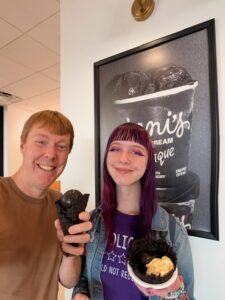 A photo of Carl and Amy standing inside Jeni's ice cream store, smiling, and holding their ice cream. Carl is in a brown shirt and has a dark brown waffle cone with dark chocolate Opaque ice cream. Amy has purple and pink hair and a purple and blue shirt. She is holding a cardboard cup with a waffle bowl and Opaque ice cream.