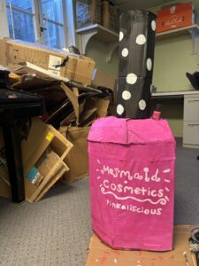 A photo of a giant nail polish bottle made out of cardboard and painted. The bottle portion is painted bright pink and has the words "mermaid cosmetics: pinkalicious." The lid is painted black with white dots. There is a tiny real nail polish bottle sitting on the cardboard one. To the left of the sculpture is a stack of unused cardboard.