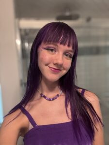 Close-up of Amy with a blurred bathroom in the background. Her hair is bright purple. Her hair is long and straight and she has bangs. She is smiling with her mouth closed and wearing a purple sundress with spaghetti straps. She is also wearing a purple beaded necklace. 