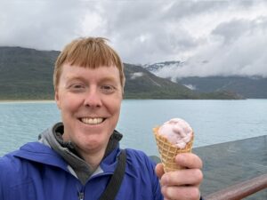 Carl smiling while holding a strawberry ice cream cone. There are mountains and an ocean behind him. He is wearing a blue jacket.