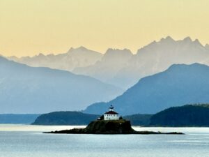 This photo looks like a painting. The colors are muted. The sky looks yellow with gray mountains in the farthest distance and then each mountain range closer gets darker. There is a lighthouse on a small island in the foreground in the middle of the ocean.