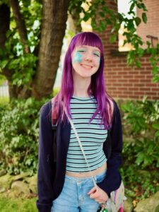 A photo of Amy standing outside with green rhododendron bushes and a tree trunk behind her. There is also a red-brick porch wall behind her. She has bright purple hair, a turquoise and navy blue striped tank top, a dark blue cardigan, and jean shorts. She is smiling. She has turquoise flowers painted on her cheeks. 