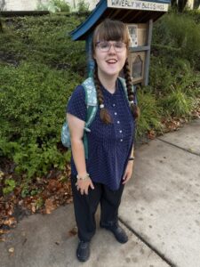 Sarah is standing in front of green bush with her feet on a sidewalk. She is wearing a blue short-sleeved blouse with little white polka dots and a row of six white buttons from the neck down. Her teal backpack is on her shoulders. She is wearing glasses and her hair is in two braids. she is smiling. 