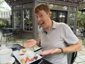 A photo of Carl in a gray polo-shirt with his hands raised to indicate a plate that has a strawberry, two grapes, and a bite of a scone and a macaron left. He is smiling in a silly way, with his eyes wide as he shows off his plate. He is outside. Behind him is a gray cement building with windows taking up most of every wall.