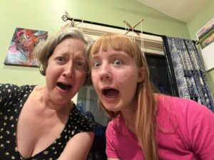 Close-up of Jenny in a black v-neck flowered short-sleeved shirt; Amy in a pink t-shirt. Their heads are together as they look at something not pictured. Their mouths are open in mock horror and shock. There is a green wall and blue curtain behind them. They cannot believe Carl did not eat any desserts as they view the picture he sent of a huge dessert buffet.
