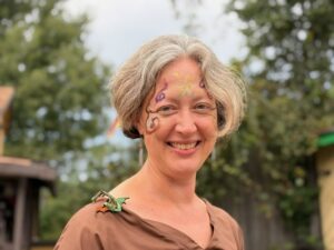 A photo of Jenny outside with blurred trees in the background. She has swirls of purple, gold, and brown facepaint on her face. She is smiling and looking slightly to her right. On her right shoulder she has a small dragon made out of leather clipped to her dress.
