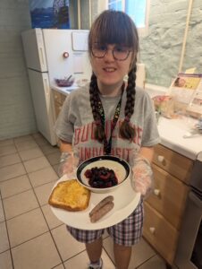 A photo of Sarah in a gray Duquesne shirt and plaid shorts, standing in an apartment kitchen with beige appliances. She is holding a plate with two pieces of sausage, a bowl of mixed berries, and a piece of French toast. Her hair is in two braids and she is wearing glasses. 