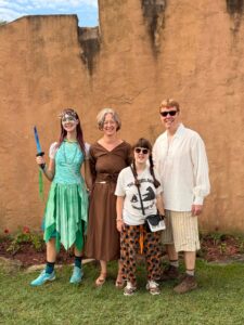 A photo of Amy, Jenny, Sarah, and Carl standing outside a brown cement wall at the Renaissance festival. Amy looks like a fairy warrior with a green skirt, turquoise corset top, a spiky crown and shell necklace. She is holding a small blue wooden dagger. Jenny is wearing a plain brown peasant dress. Sarah is wearing a white t-shirt with a black bear on it and halloween pajama pants with cats and pumpkins. Carl is wearing a loose white shirt and loose beige shorts that look like what a tradesman in the renaissance might wear.