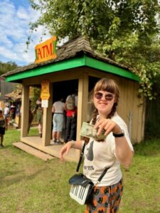 A photo of Sarah in sunglasses, a white t-shirt with a black bear on it, and halloween pajama pants with cats and pumpkins. She is standing outside in the sunshine in front of the small ATM booth at the Renaissance festival. She is holding up a $20 and smiling.