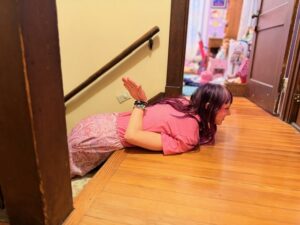 A photo of Amy dressed in a pink skirt and pink shirt, lying on her belly on the top steps and landing of the stairs next to her room. She is looking ahead with her head up and her right arm is behind her back with her hand vertically pointing towards the ceiling to be a fin