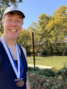 A photo of Carl standing outside in the sunshine with trees and grass behind him. He is wearing his rowing race uniform and hat and has two medals around his neck.