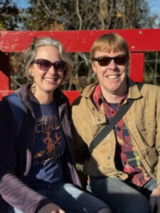 A photo of Jenny and Carl riding in a red hay wagon. They are smiling and wearing sunglasses. Jenny has a purple fleece over a blue t-shirt. Carl has a red plaid button-down shirt visible under a tan button-down coat that is unbuttoned.