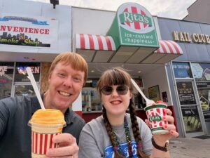 A photo of Carl and Sarah smiling in the sun as they hold their frozen treats from Rita's. You can see Rita's in the background, with the red and white striped awning and green Rita's sign.