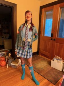A photo of Amy standing inside next to our front door. She is wearing a blue and white plaid skirt, a light blue sweater vest, a jean jacket, and a pink and blue tie.