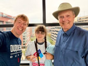 A photo of Carl, Sarah, and Grandpa in front of a sunny window. Carl is wearing a dark blue Duquesne shirt. Sarah is wearing a white t-shirt with some colored squares on the front, and Grandpa is wearing a sun hat and long-sleeved blue button-down shirt.