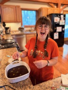 Photo of Sarah standing at a kitchen counter by a small white casserole dish with brownies. She is wearing an orange t-shirt with Peanuts characters. Her hair is in two braids and she has an enormous smile.