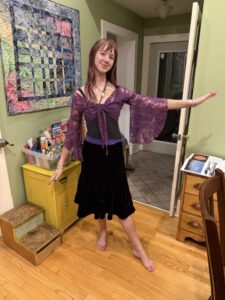 Photo of Amy standing next to a small yellow dresser and a small pine dresser. The floor is hardwood. There is an abstract colorful quilt on the green-painted wall to Amy's right. She is sort of in a doorway. She has her hair long and free and is wearing a knee-length velvety black skirt and a black top under a lacy purple sweaterlet with drapey bell-like sleeves.