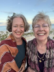 A photo of Jenny and her mom, Janet, outside and smiling with their hair blowing in the wind. Behind them you can see traces of sand and water at the beach. Jenny has a teal top. and an orange and red shawl. Janet has a purple top and a purple and white decorative scarf.